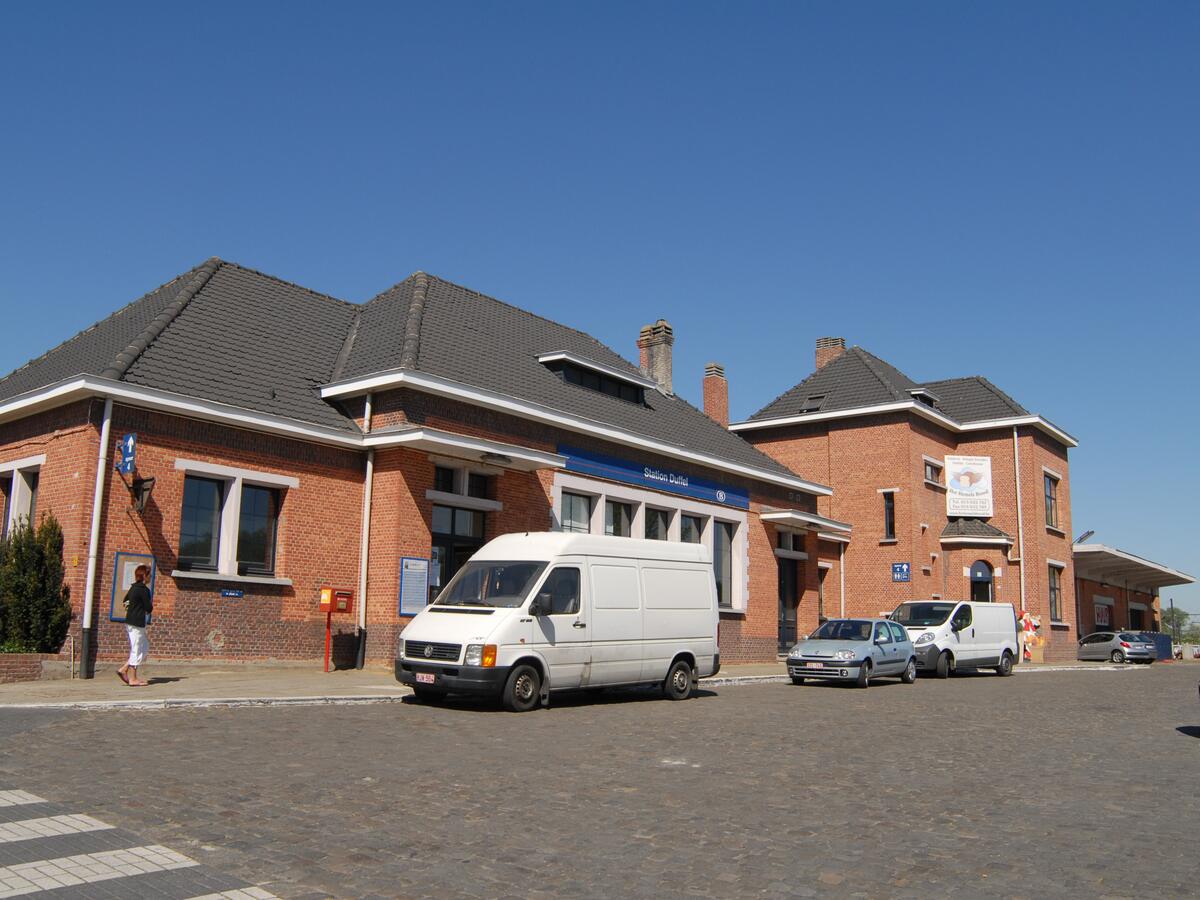Het stationsplein van Duffel in de zon, met het stationsgebouw en een paar geparkeerde bestelwagens